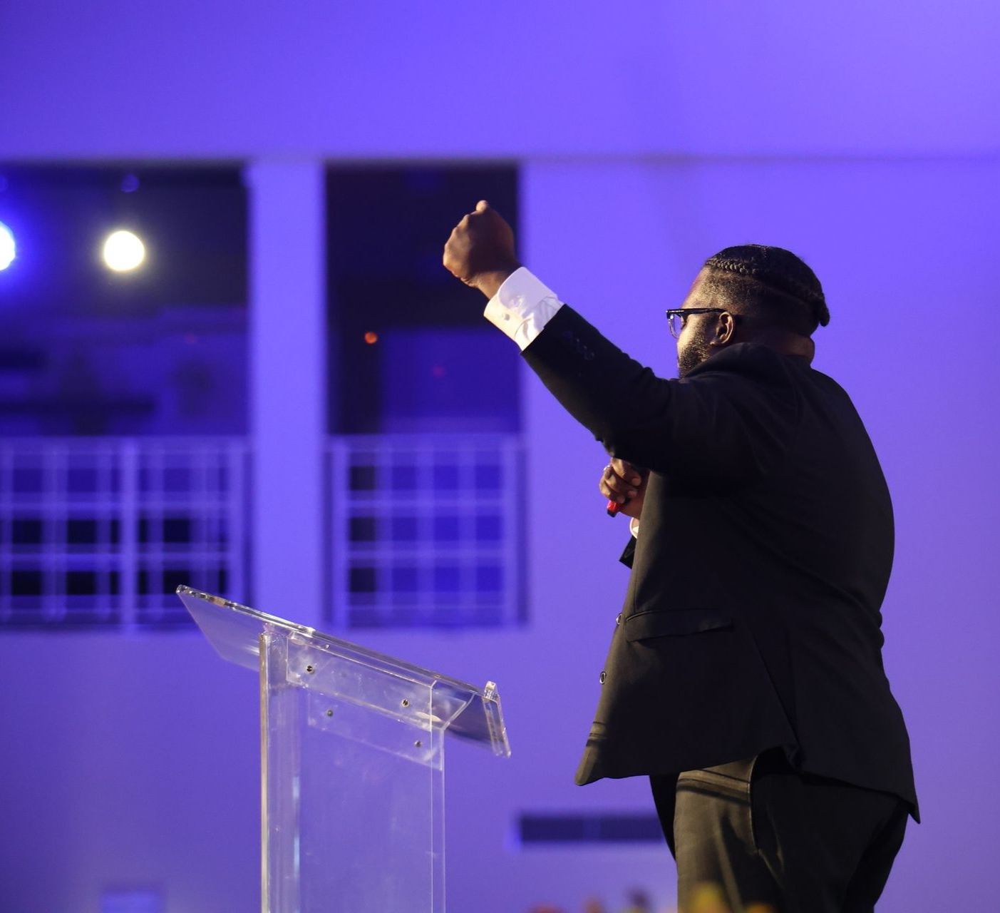 Speaker in a dark suit raises a fist behind a clear podium under purple stage lighting.