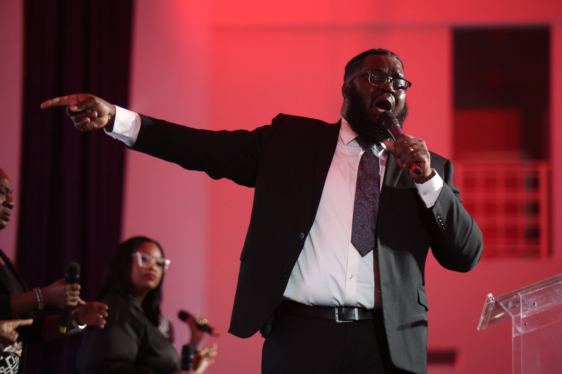 Man in suit speaking into a microphone and pointing on a pink-lit stage