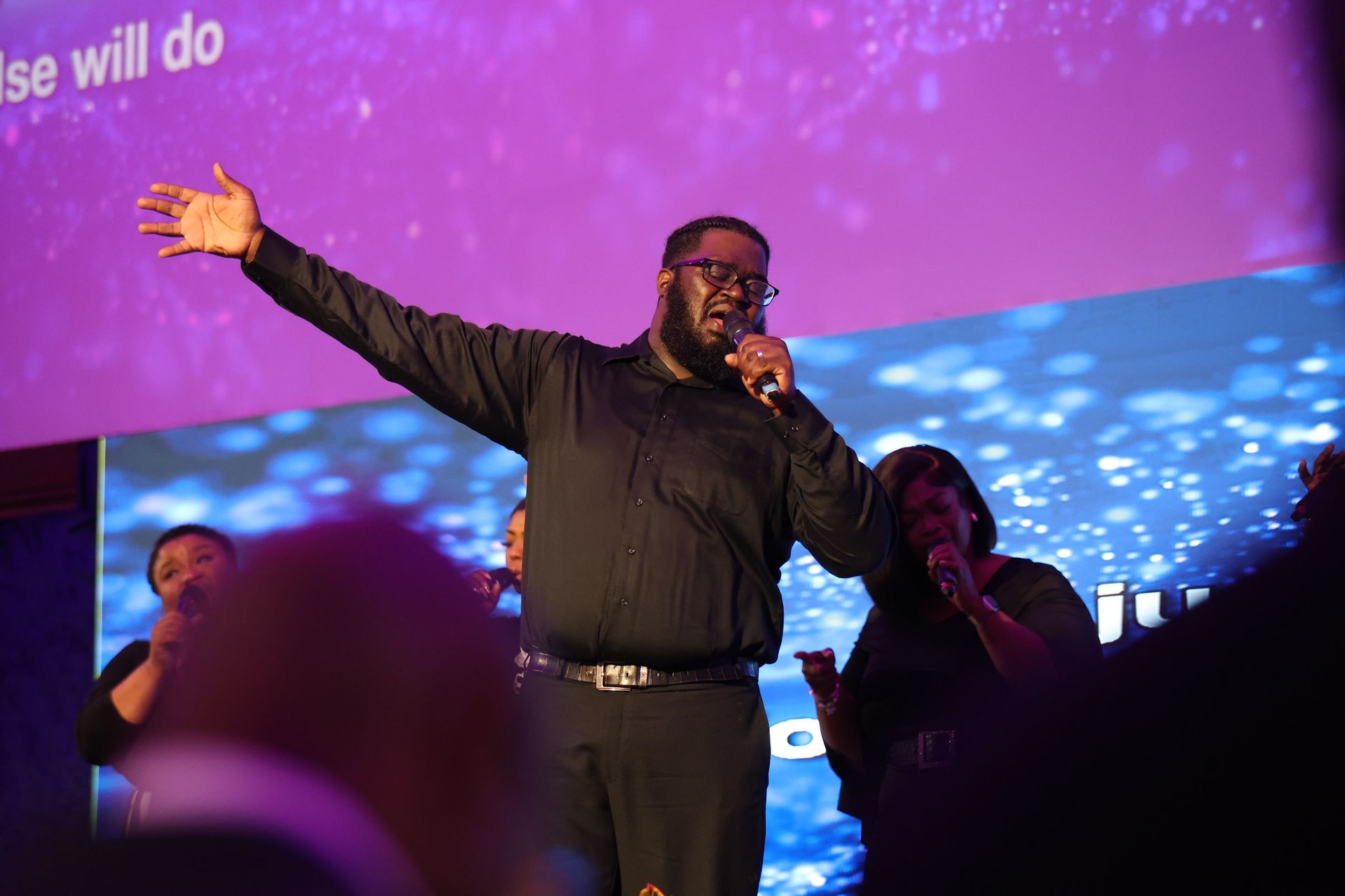 Man singing on stage with one arm raised, lit by purple and blue concert lights.
