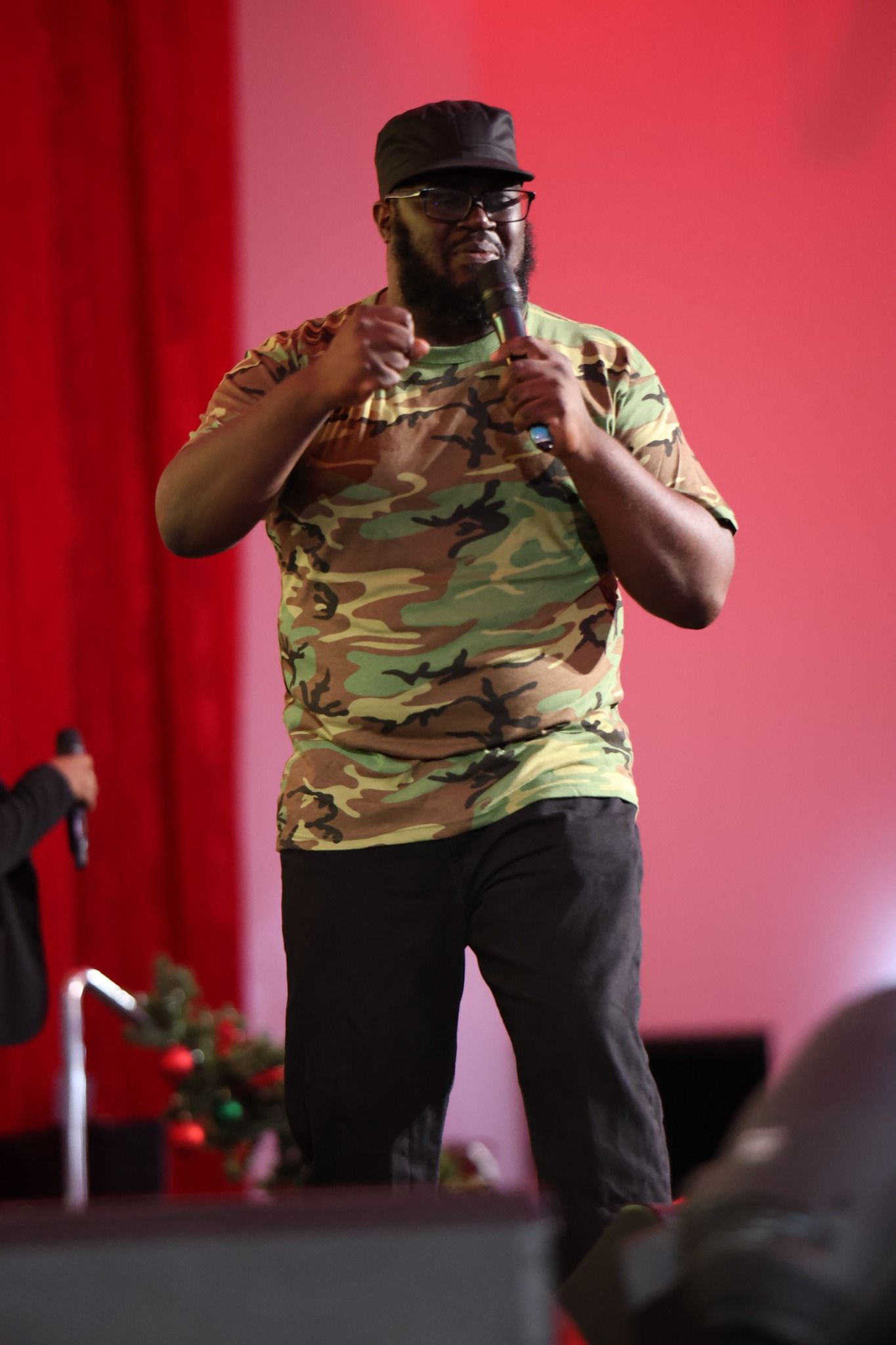 Speaker on stage in camouflage shirt and black cap, gesturing against a red backdrop.