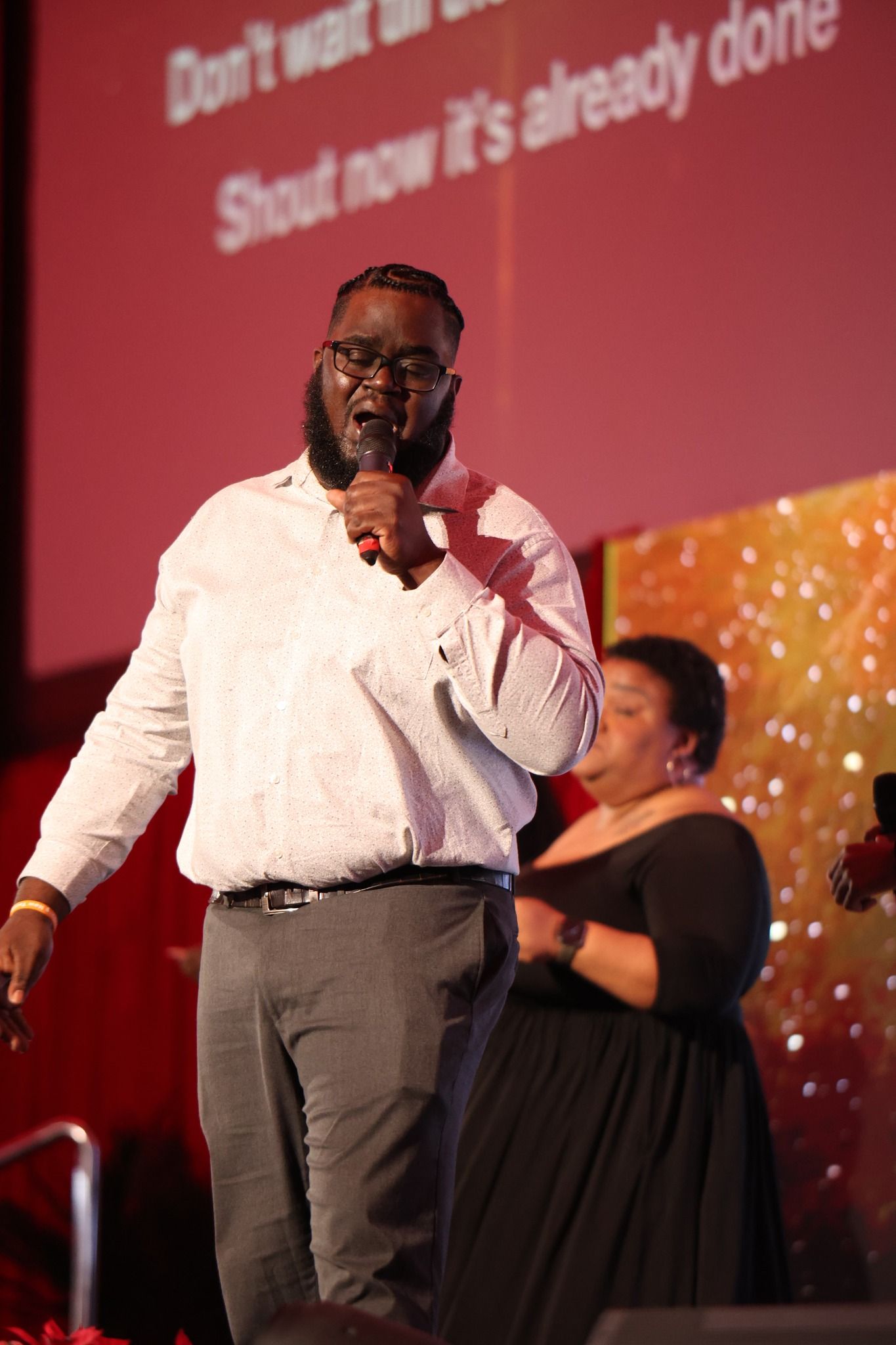 Man speaking into a microphone on stage, with a seated woman and a red backdrop behind him.