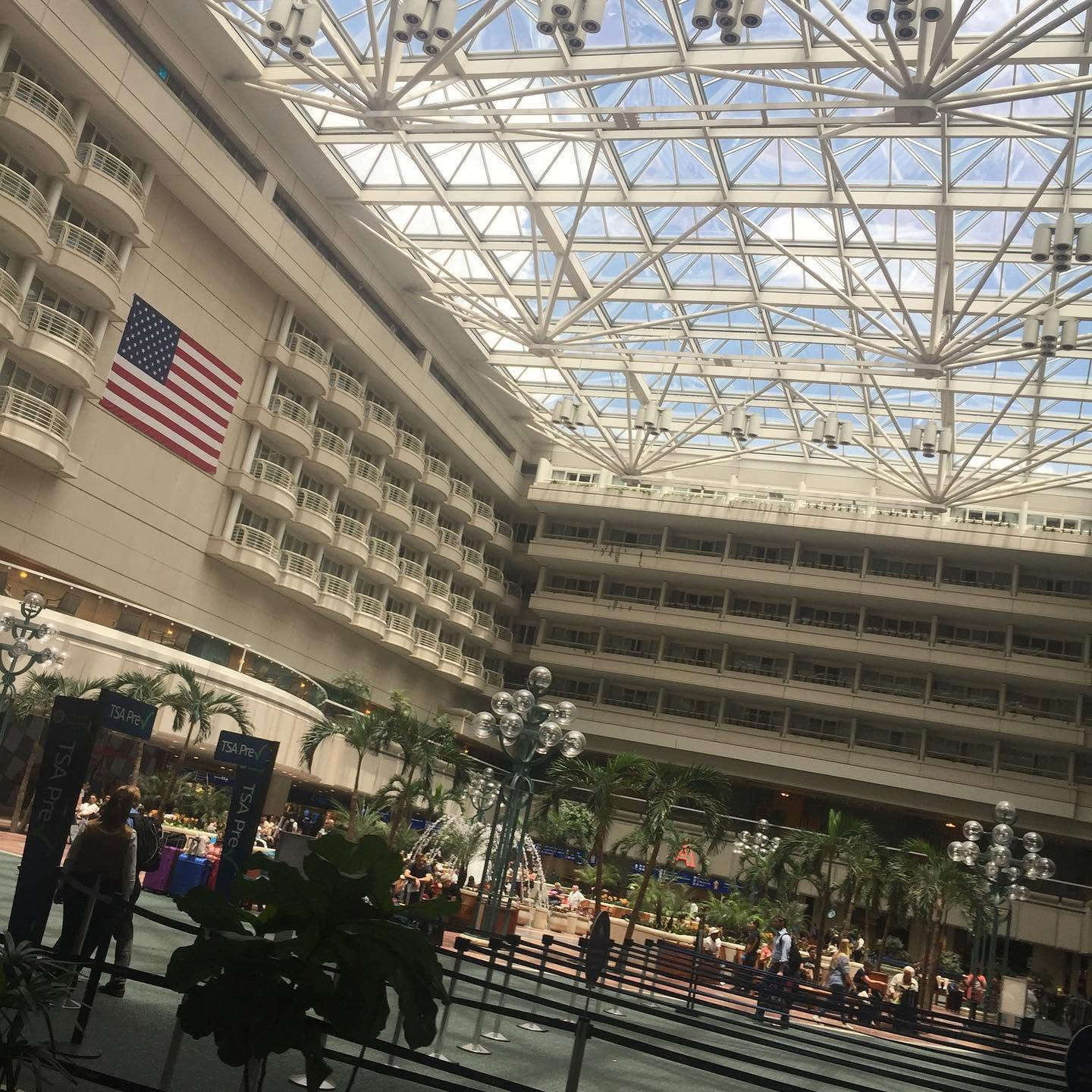 Large atrium with a glass ceiling, American flag, and people gathered around indoor greenery and lights