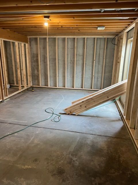 Basement with wooden wall framing, concrete floor, and stacked lumber; construction in progress.
