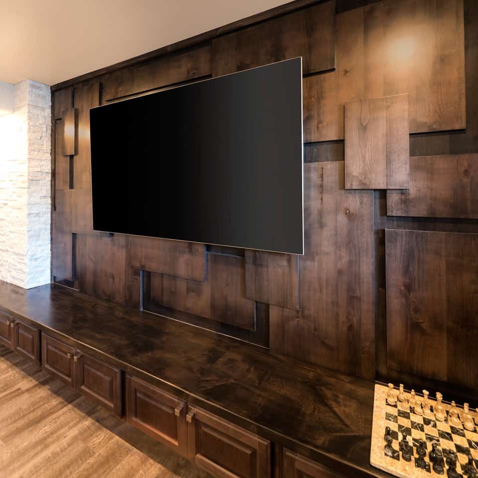 Large TV mounted on a dark wood wall with built-in storage, next to a stone wall and a chess board.