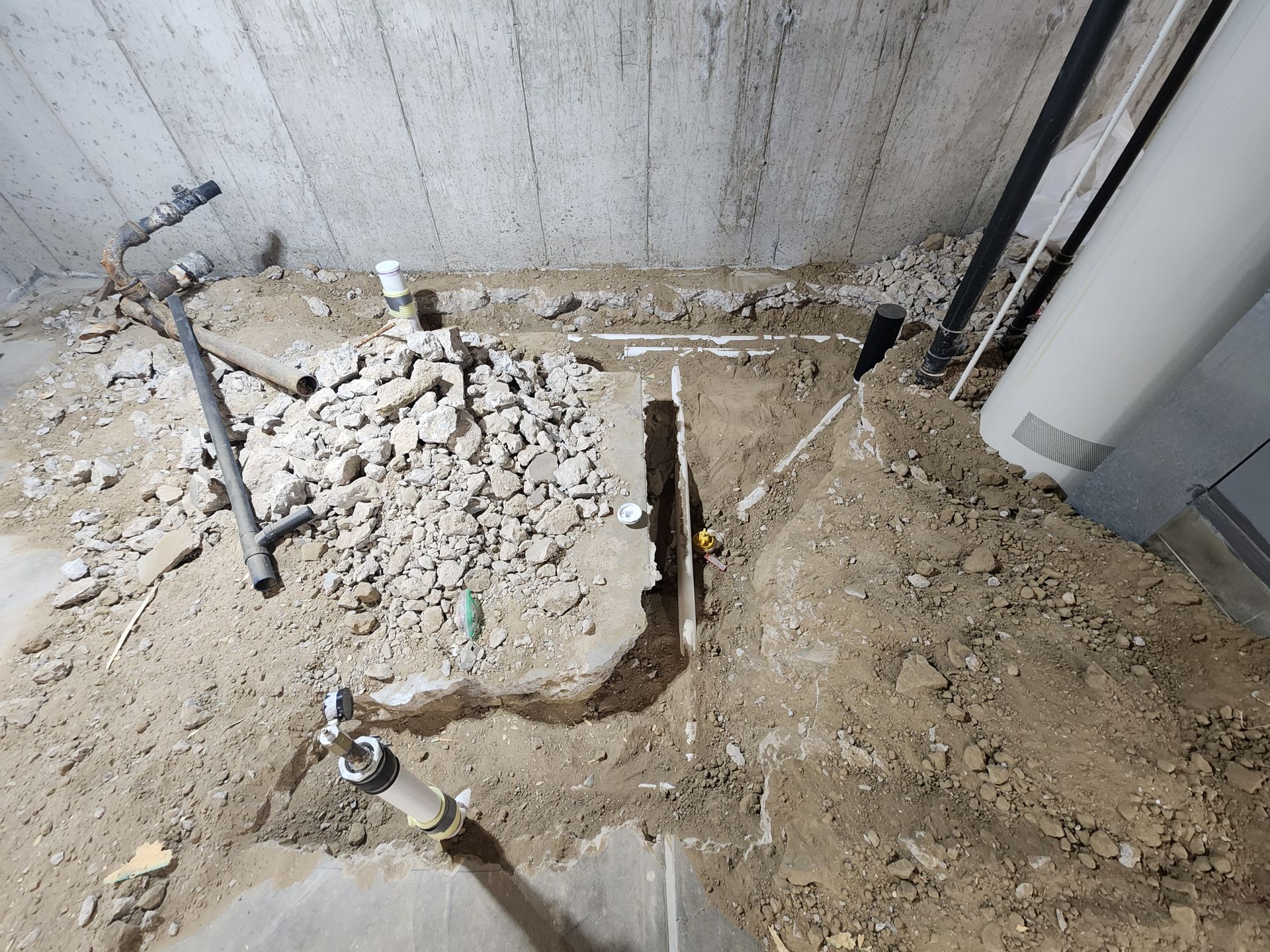 Construction site with exposed pipes and debris in a trench under a concrete ceiling.