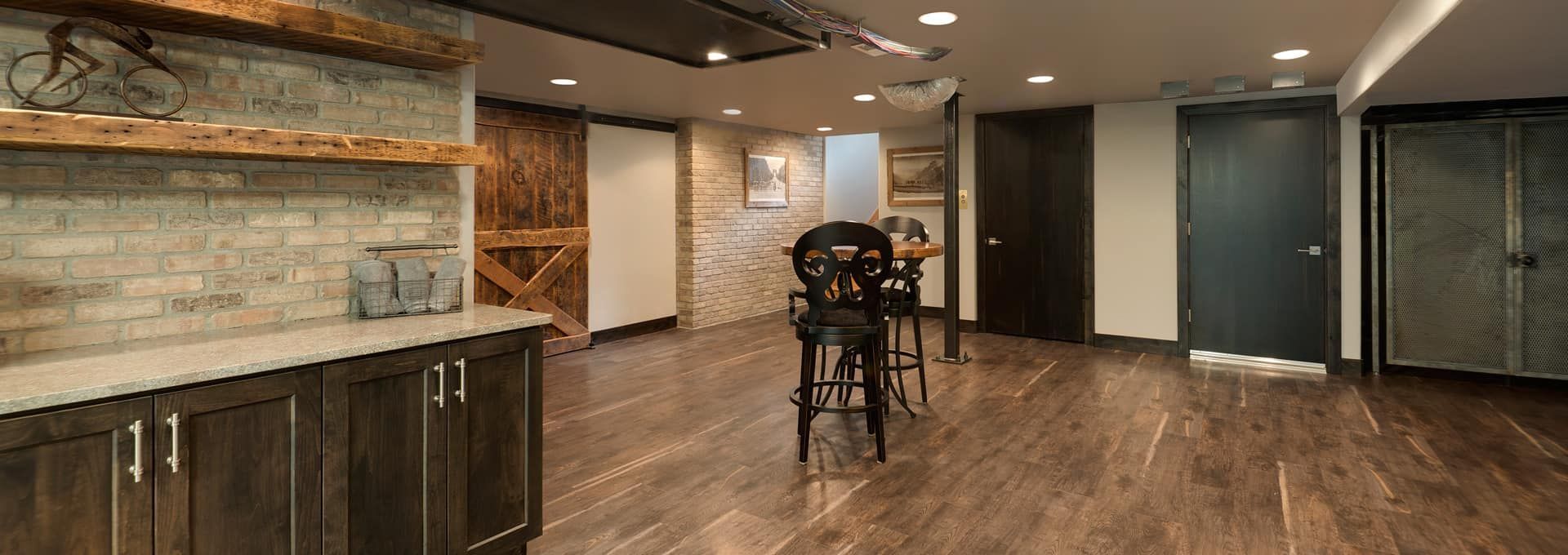Rustic basement with brick wall, wooden floor, bar stools, and dark cabinetry.