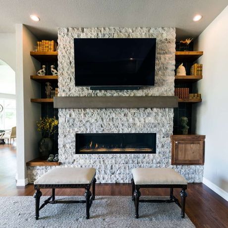 Fireplace with stone surround, TV, shelves with decor, and two upholstered ottomans in a modern living room.