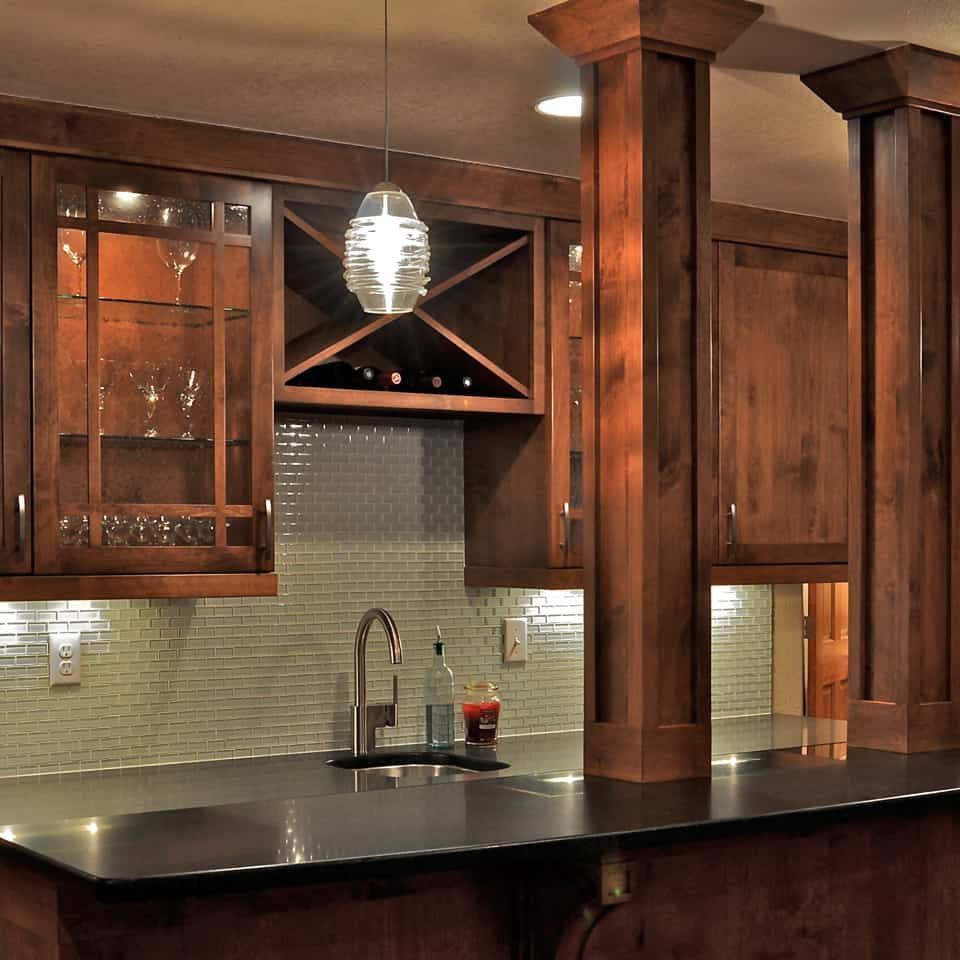 Wooden home bar with dark countertop, glass cabinets, and a sink.