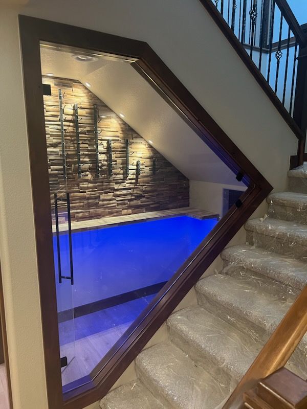 Small, blue-lit indoor pool visible through a glass wall beneath a staircase, accented by stone and wood.
