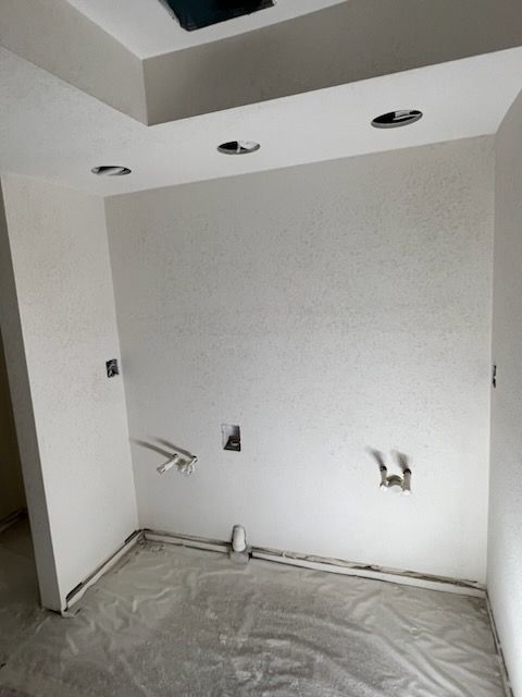Interior room under construction with white walls, ceiling, and gray flooring; electrical and plumbing fixtures visible.
