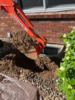 An orange excavator bucket scoops dirt near a brick building and a window well.