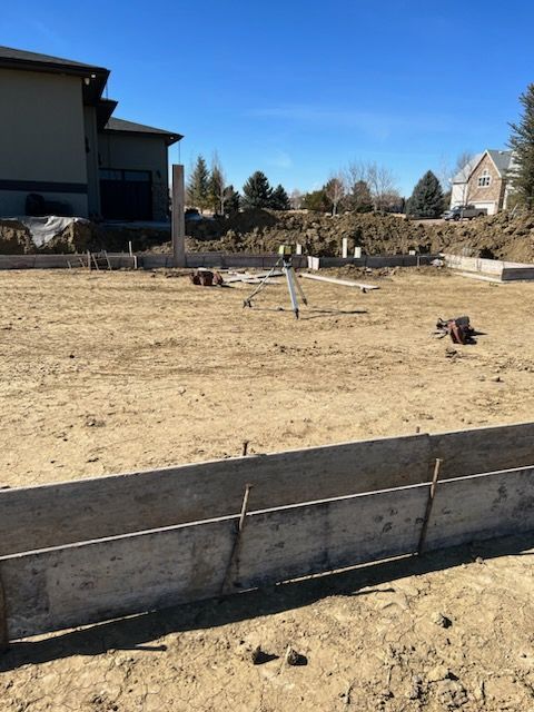 Construction site: concrete forms outlining a foundation, exposed dirt, and neighboring houses under a blue sky.