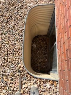 Window well with gravel and brick wall. Beige corrugated metal curves inward.  Foot and shoe visible.