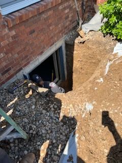 Person in a hole, next to a brick wall with a window, digging near a building.