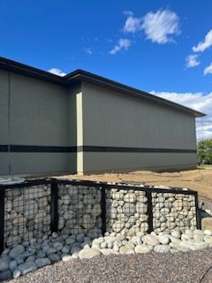 A low building with a gray exterior, black trim, and a stone retaining wall with a blue sky background.