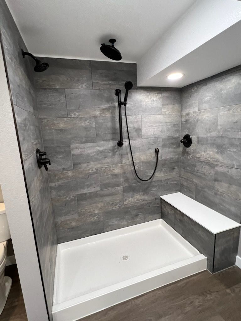Modern shower with gray tiled walls, black fixtures, white shower pan and built-in bench.