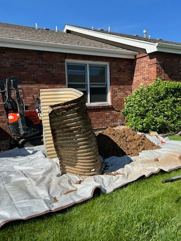 A corrugated metal window well being removed next to a brick house with a mini-excavator on a sunny day.