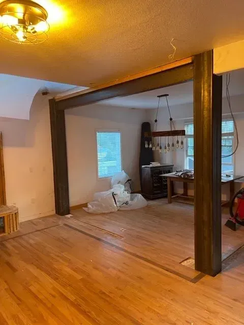 Open-concept interior with wood floors and dark brown support beams. A dining area with table and chandelier is visible.