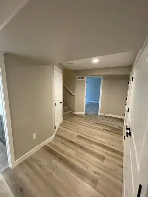 Basement hallway with wood-look flooring, tan walls, white doors, and a staircase.