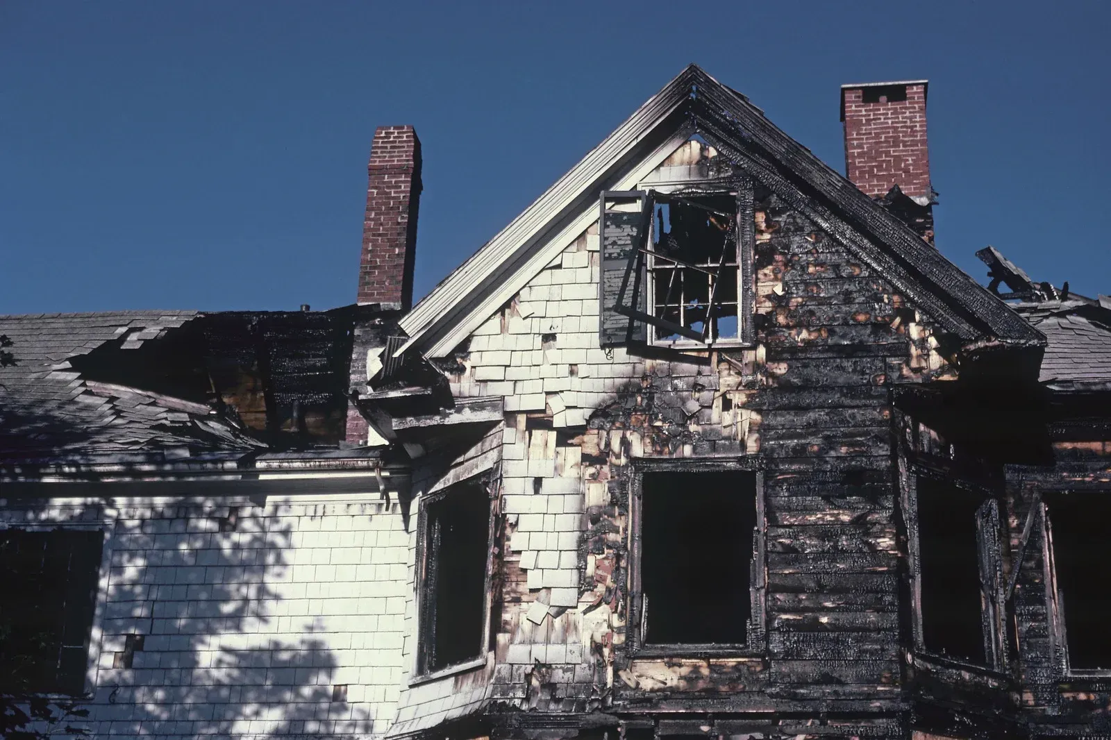 Burned-out house with charred siding, broken windows, and two brick chimneys against a blue sky.