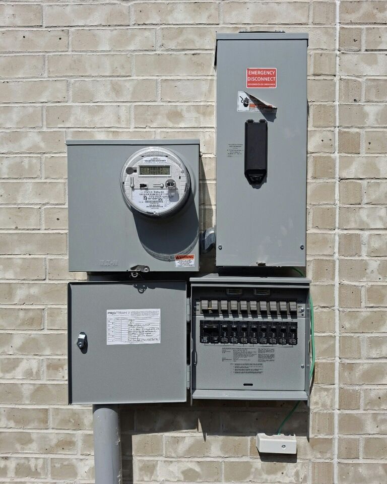Electrical panel components mounted on a brick wall: meter, panel box, breaker box, and conduit.