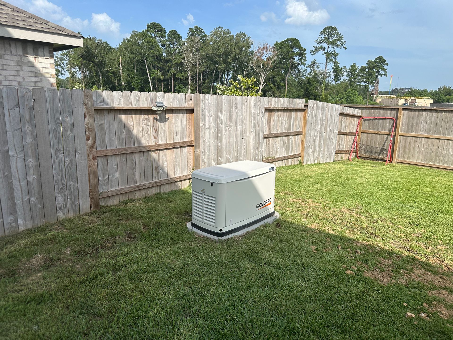 A tan home generator sits on grass in a fenced backyard on a sunny day.
