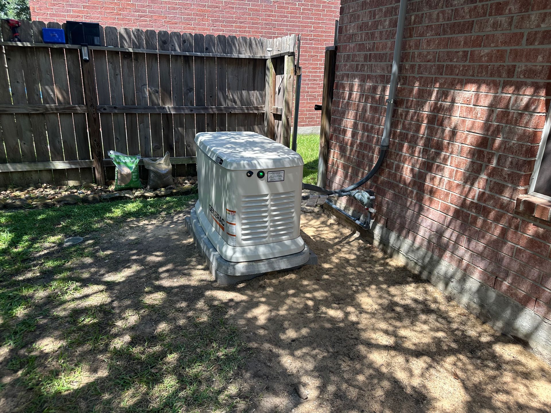 A large, beige generator sits next to a brick building and a wooden fence on grass.