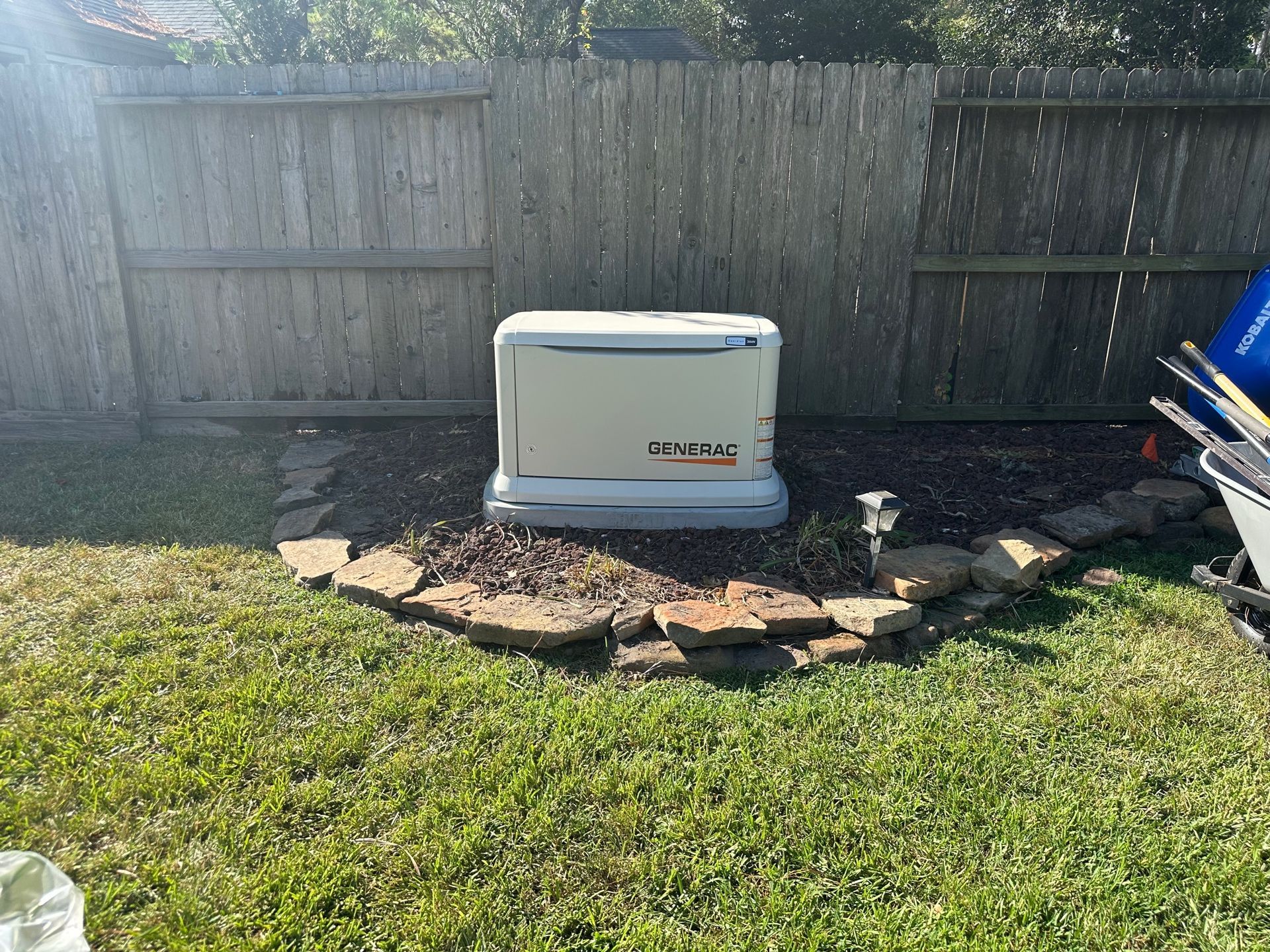 Generator in a backyard, beside a wooden fence, surrounded by landscaping rocks.