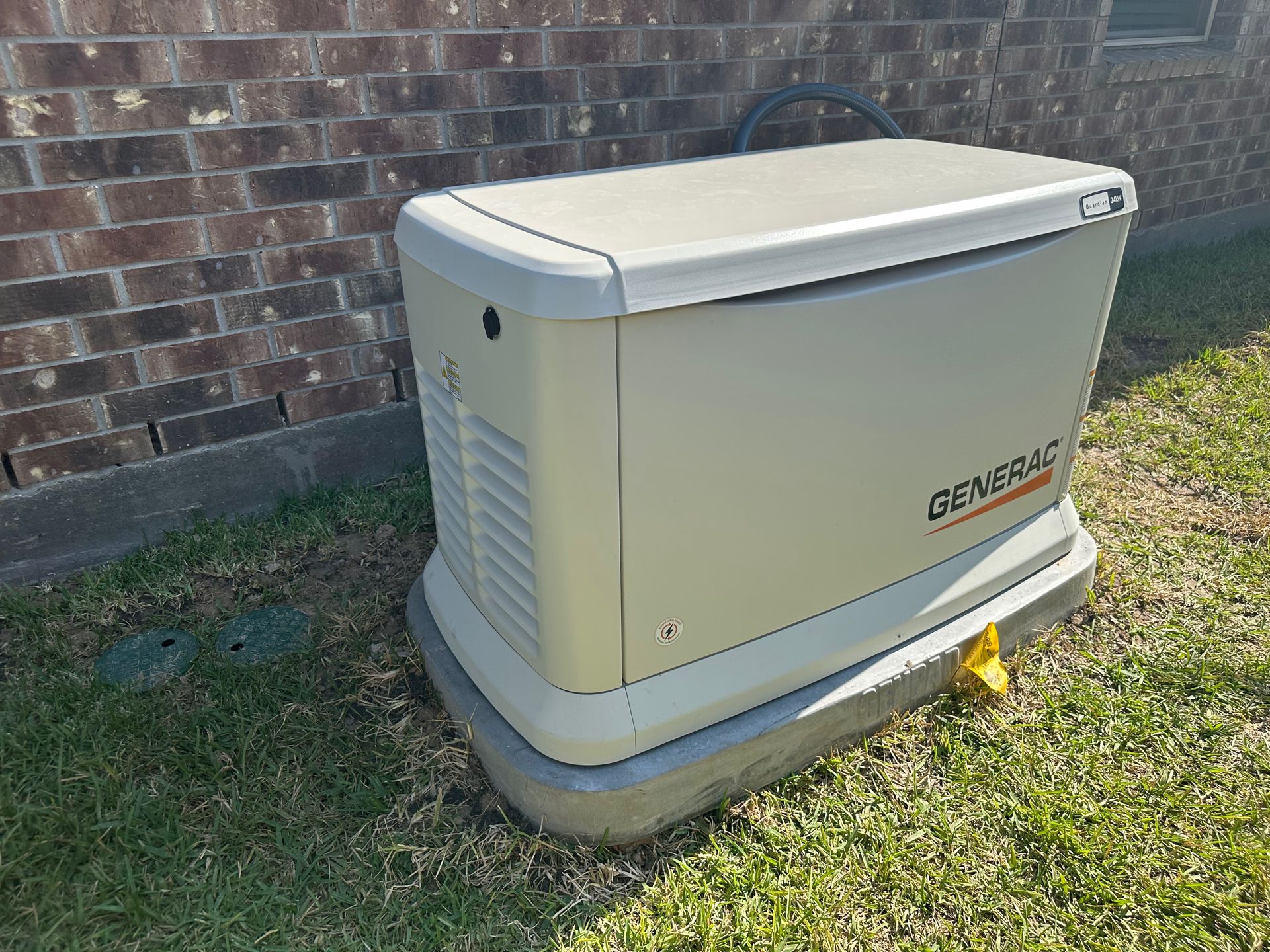 A beige Generac generator on a concrete base next to a brick wall and grass.