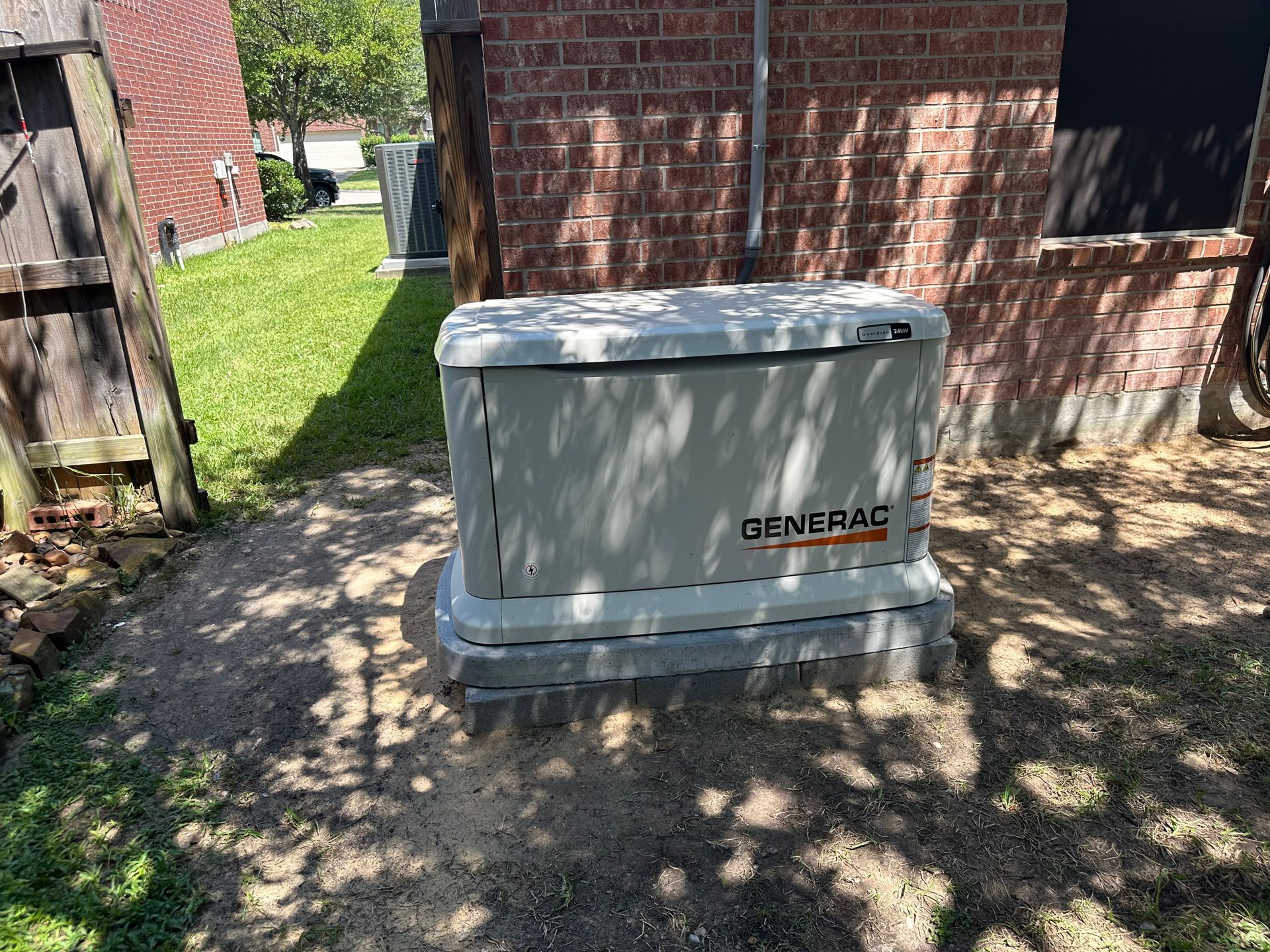 A Generac home generator on a concrete base, beside a brick wall and grass.