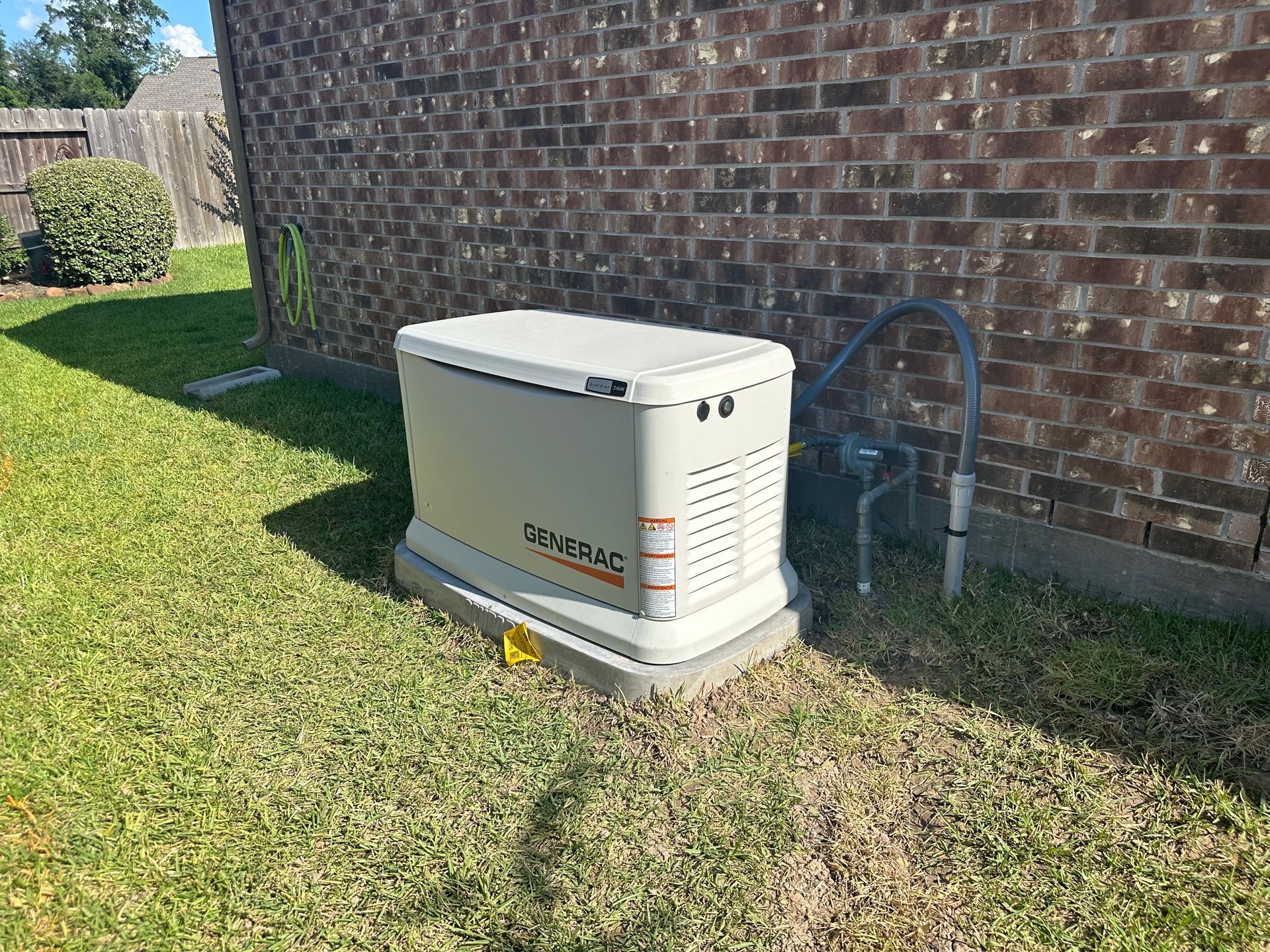 Generator in beige casing on a concrete pad, next to a brick wall and a gas pipe, in a grassy yard.