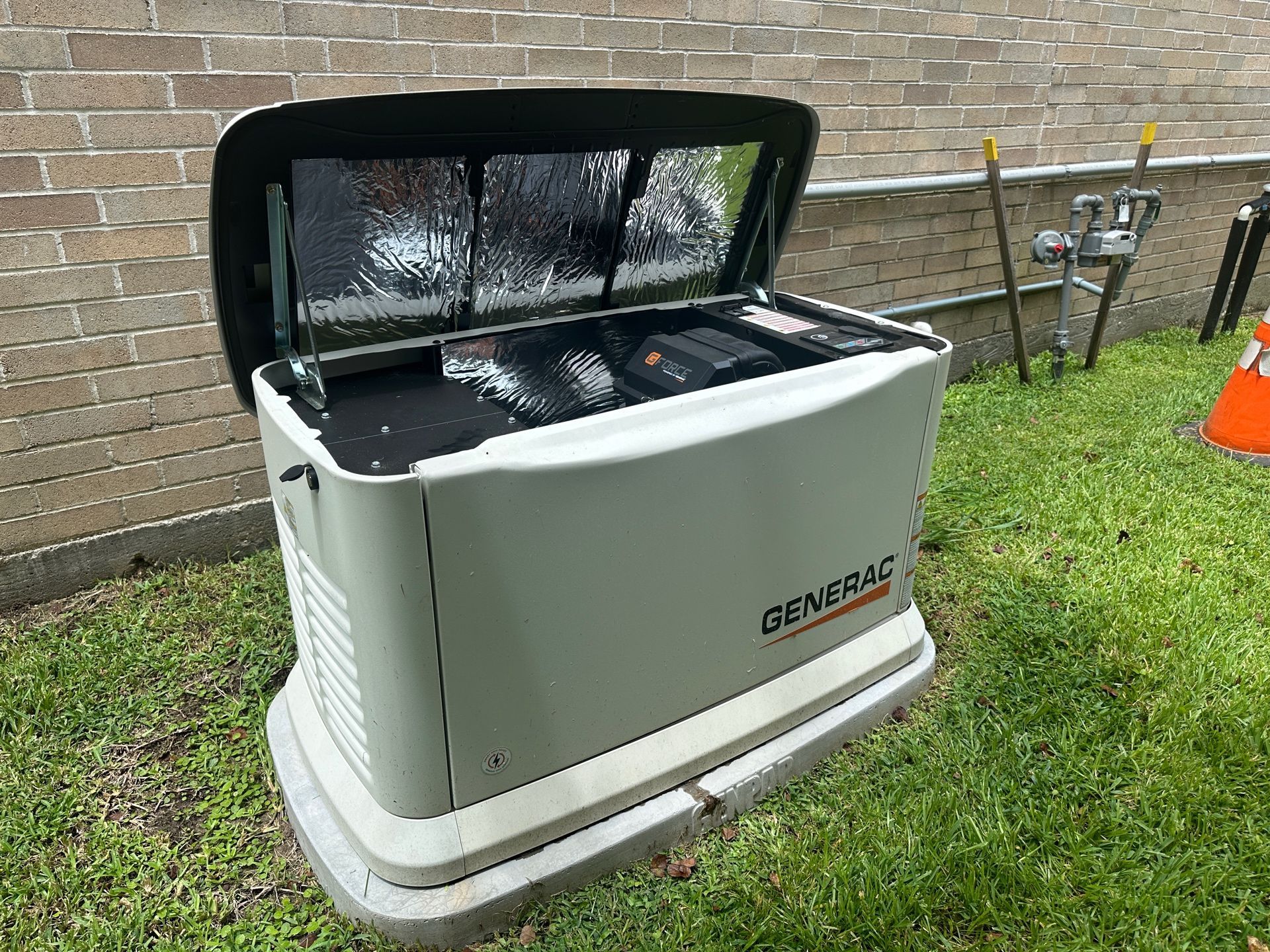 Generac home generator with open lid in a yard, next to a brick wall.