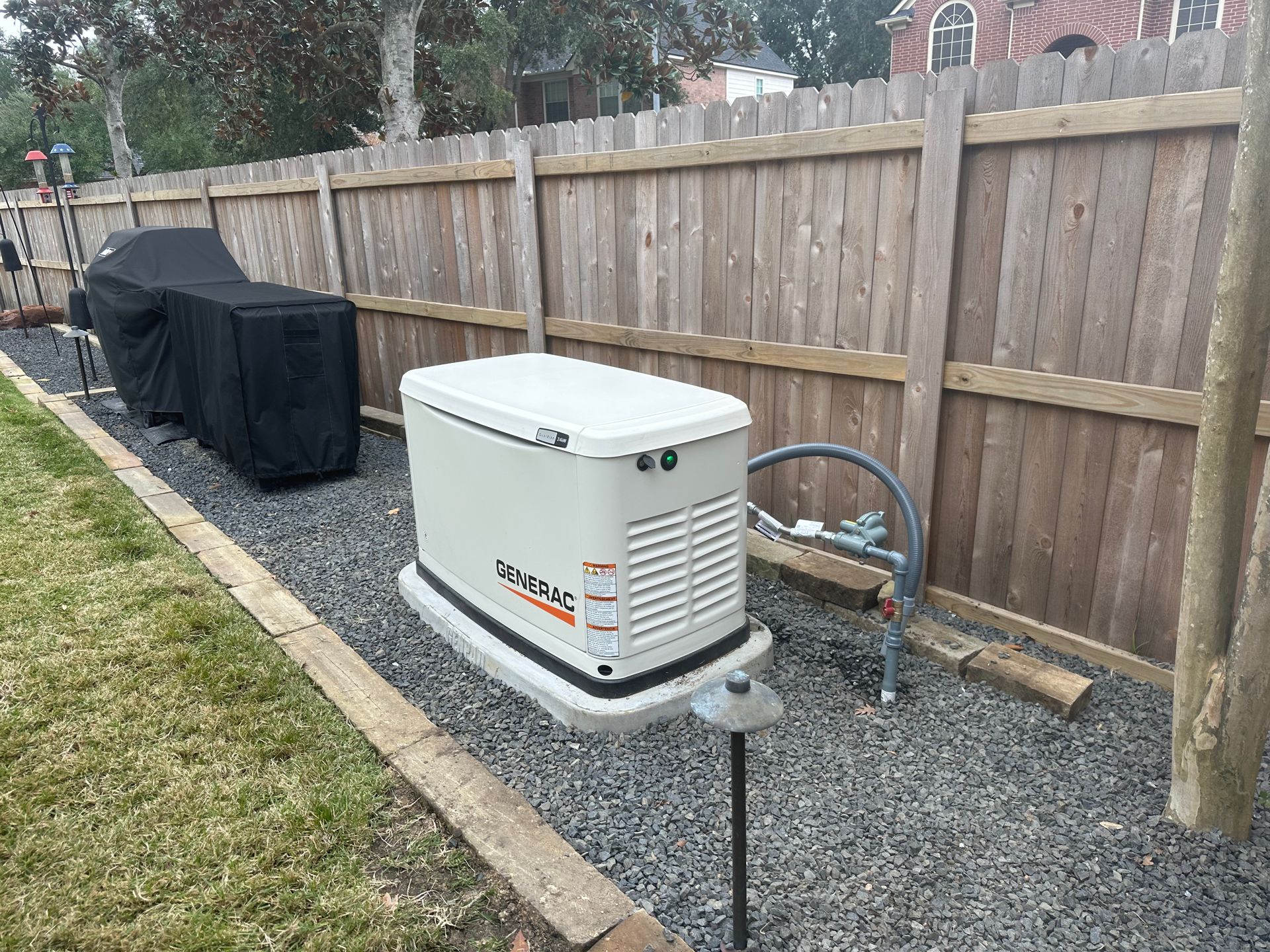 Generator next to wooden fence and covered grill, on gravel next to a lawn.