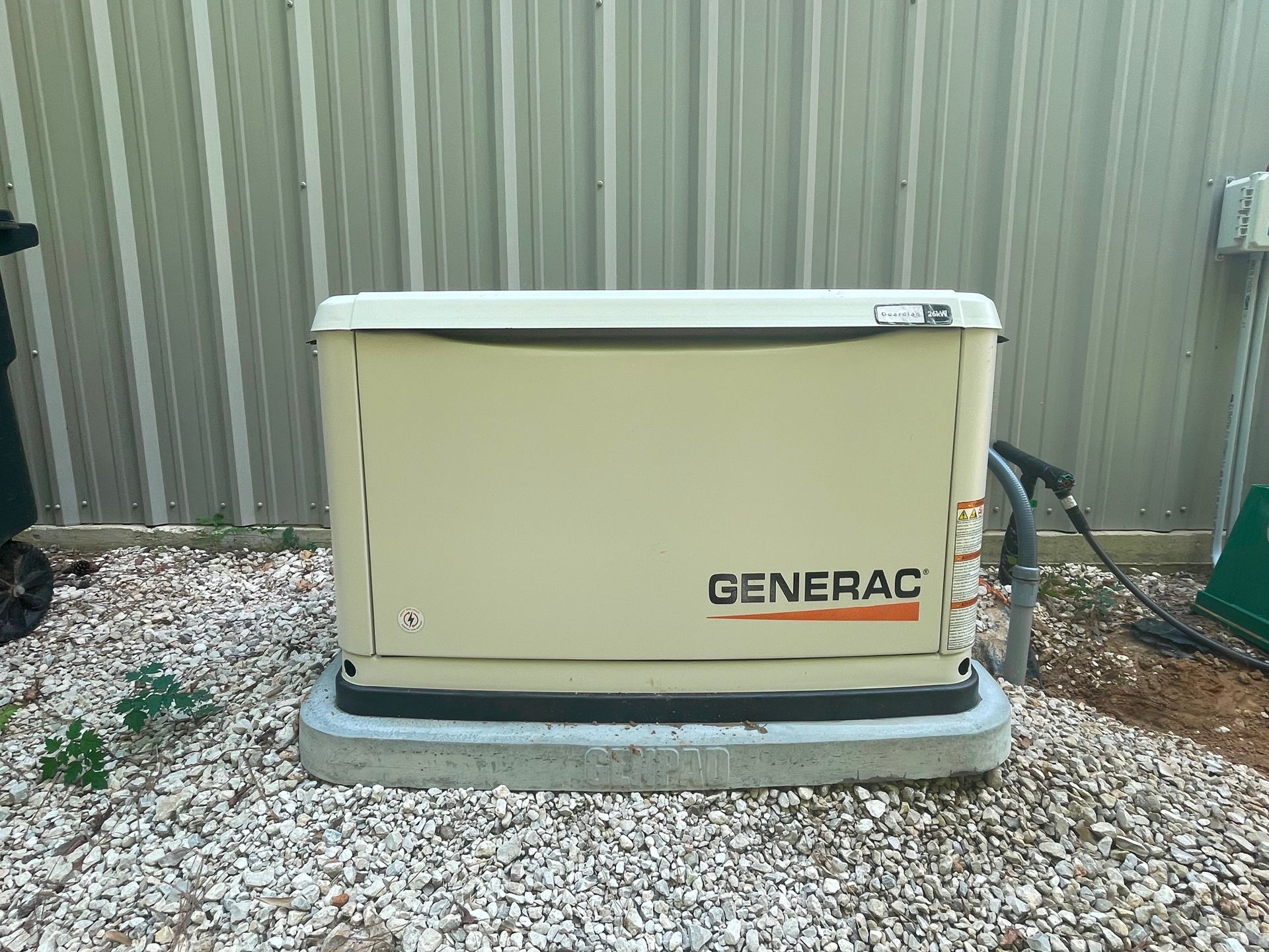 Tan Generac generator on concrete base in a yard with gravel. Gray metal siding background.