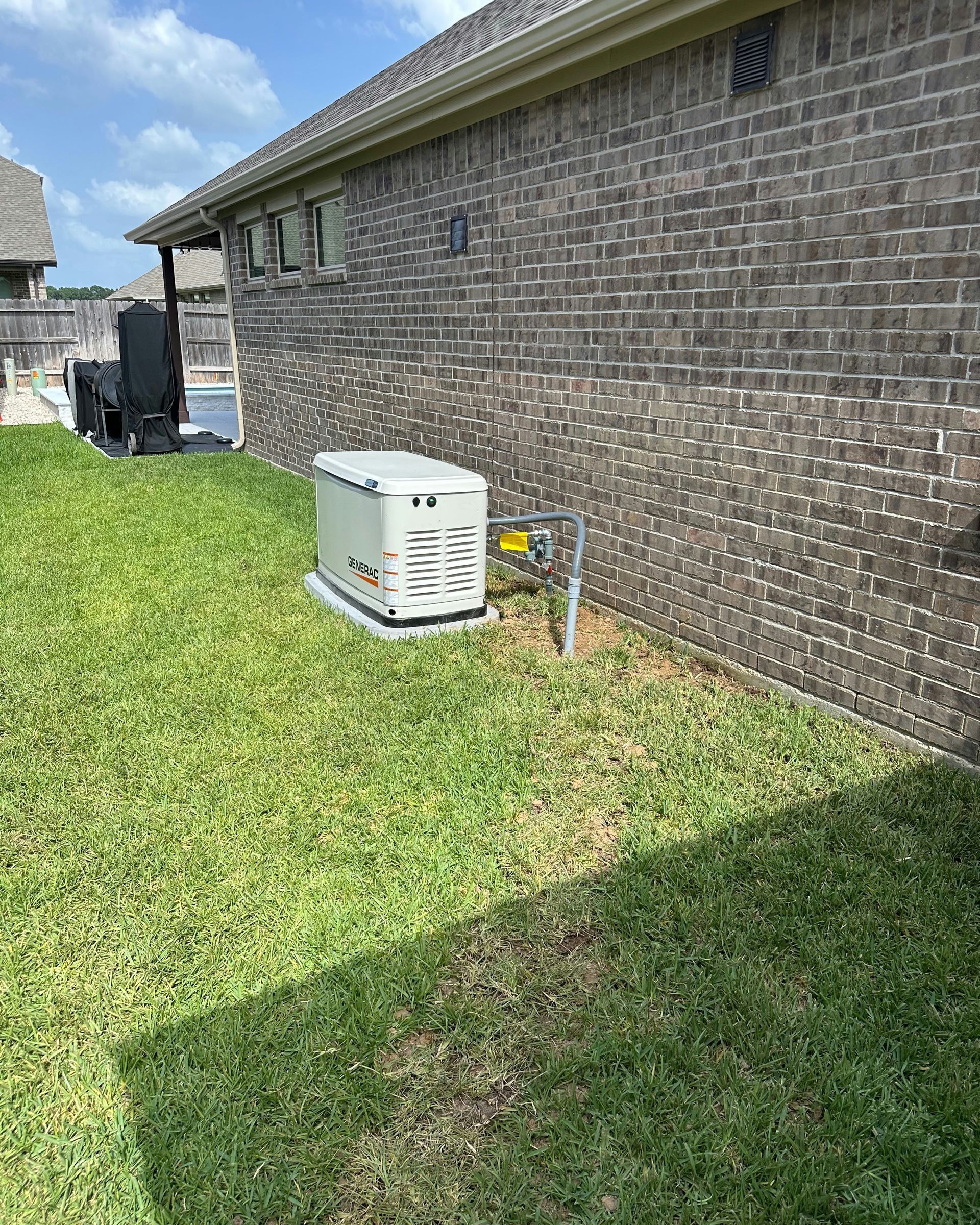 A residential standby generator sits on a concrete pad near a brick house, connected with metal conduit.