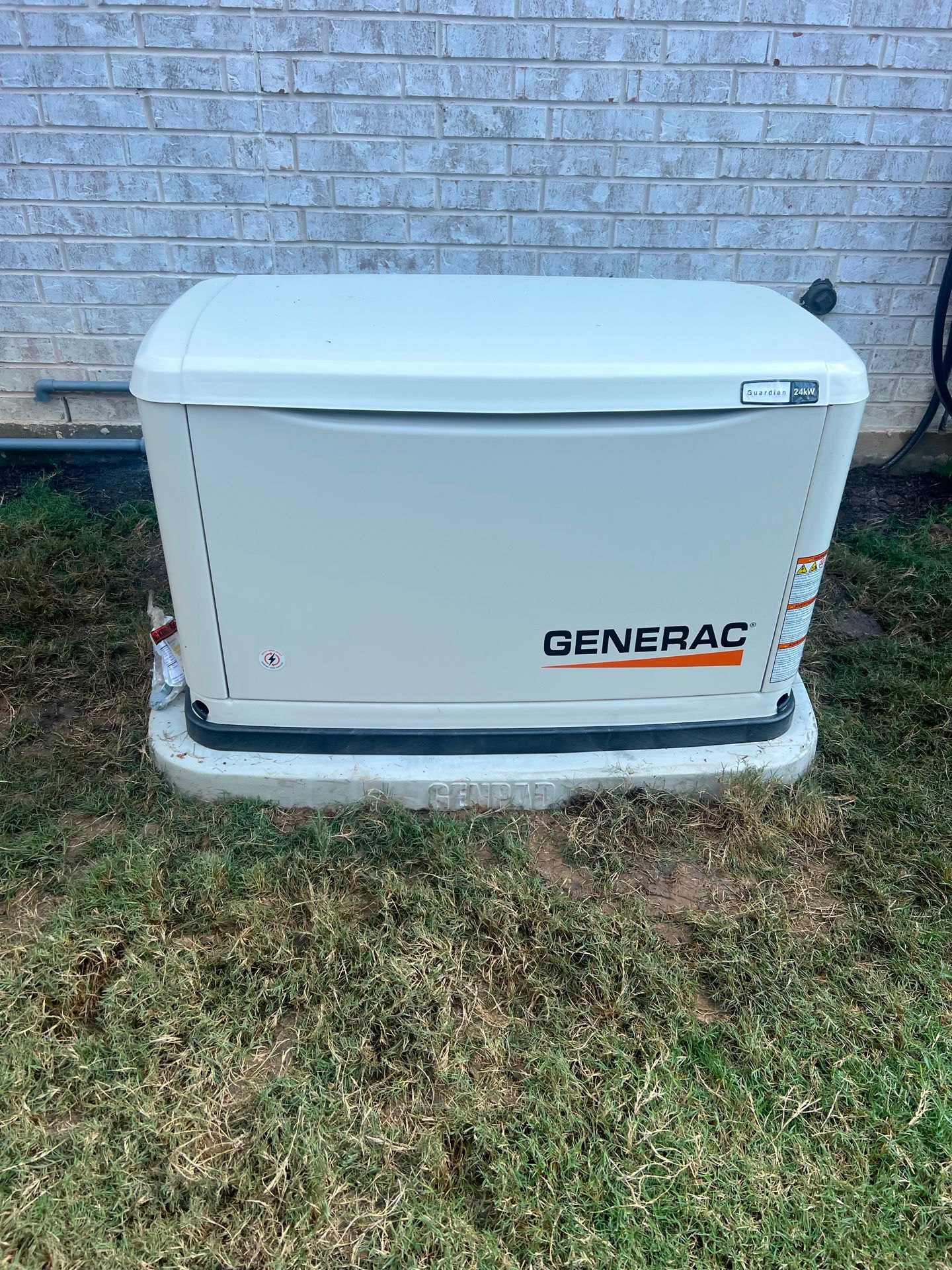 A Generac home generator on a concrete pad, against a brick wall, in a grassy yard.