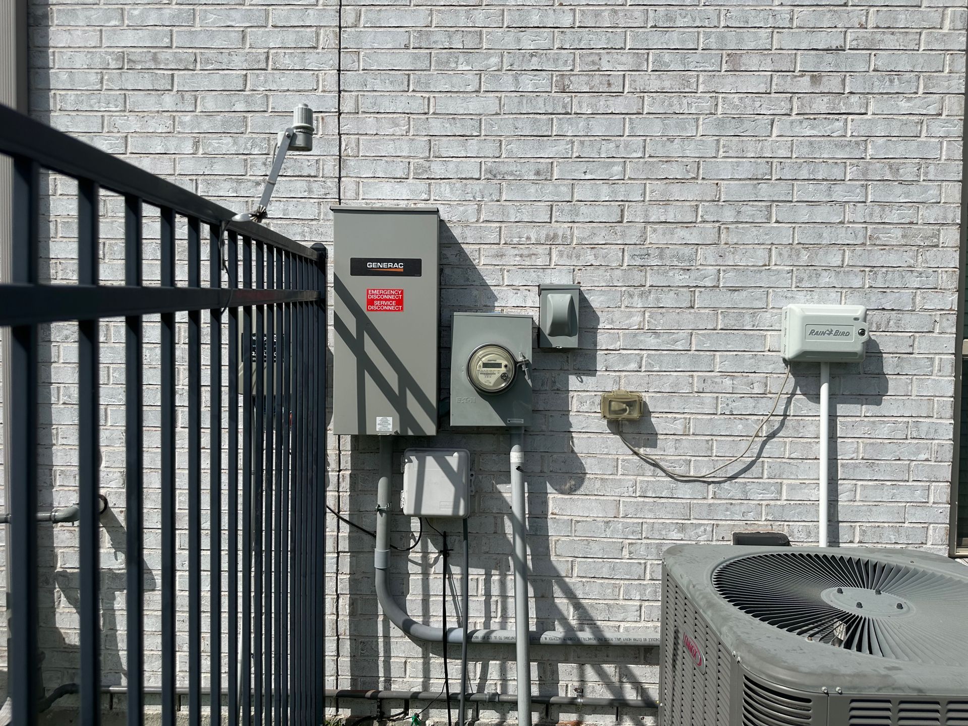 Exterior electrical panel, meter, and air conditioning unit on a brick wall next to a black metal fence.
