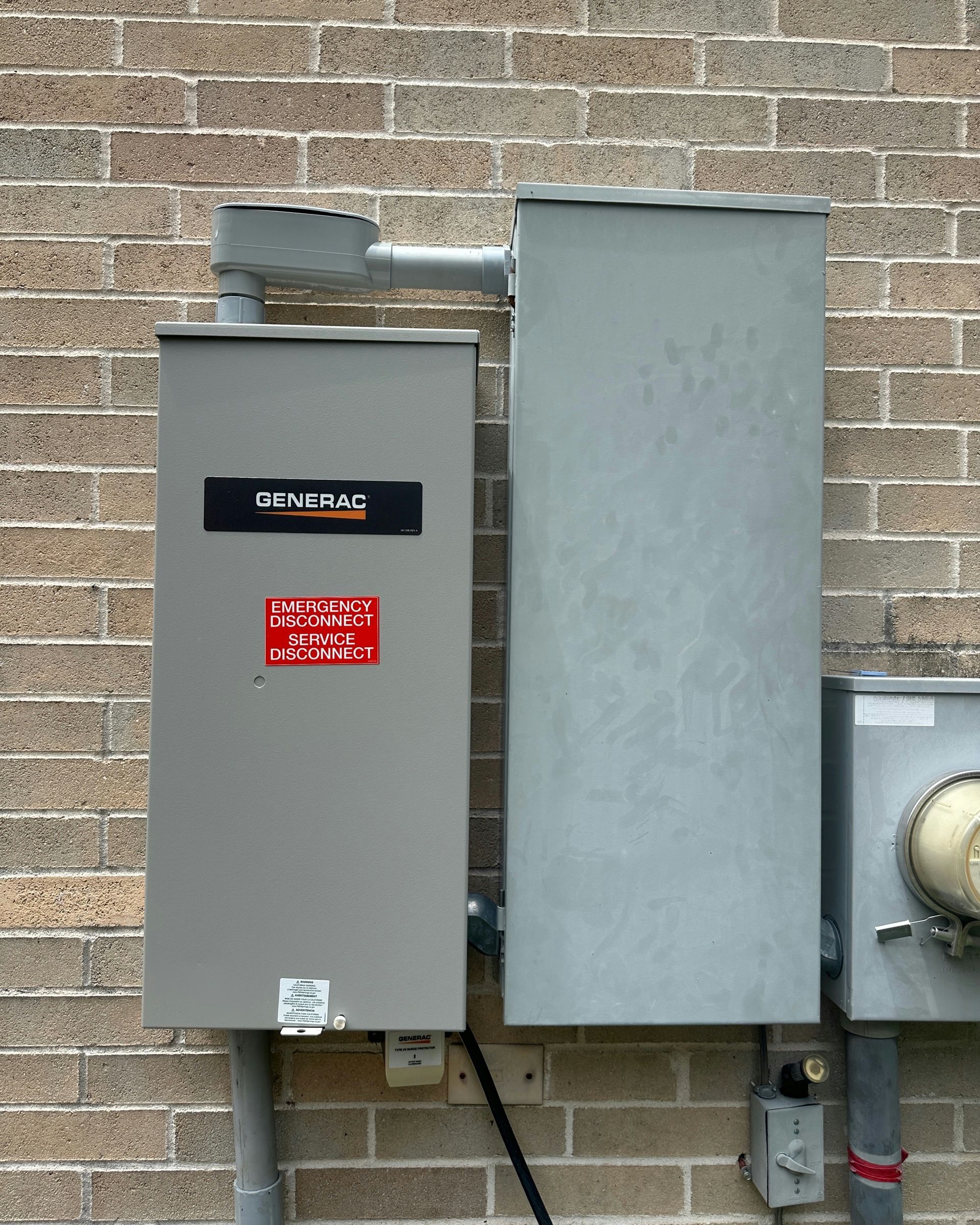 Gray electrical generator box and panel mounted on a brick wall with conduit and wiring.