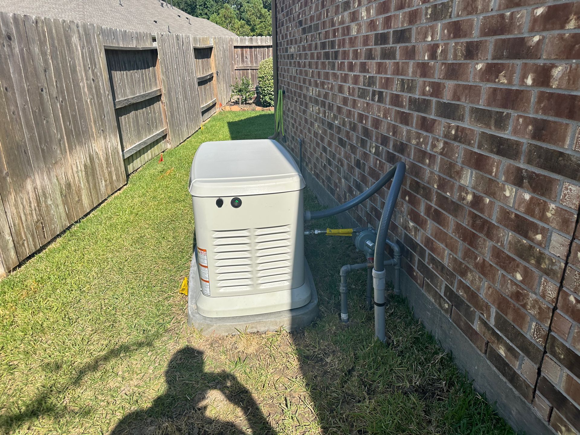 Generator next to a brick wall and wooden fence, set in a grassy yard.