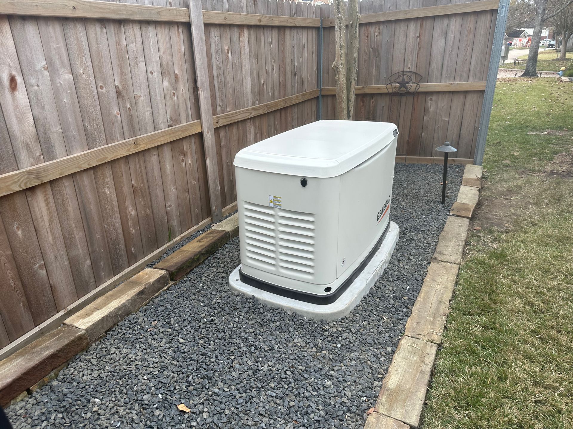 Gray standby generator in a gravel area beside a wooden fence.