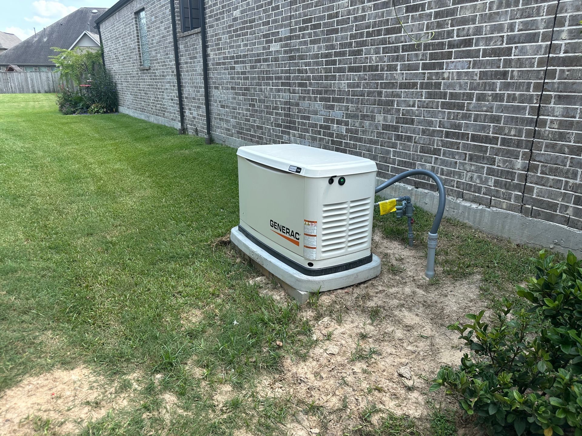 A standby generator is on a concrete pad next to a brick house on a grassy lawn.
