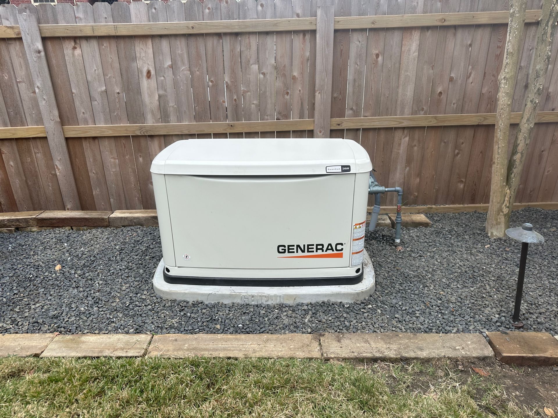 Generac generator in a gravel-filled yard, next to a wooden fence and small light fixture.