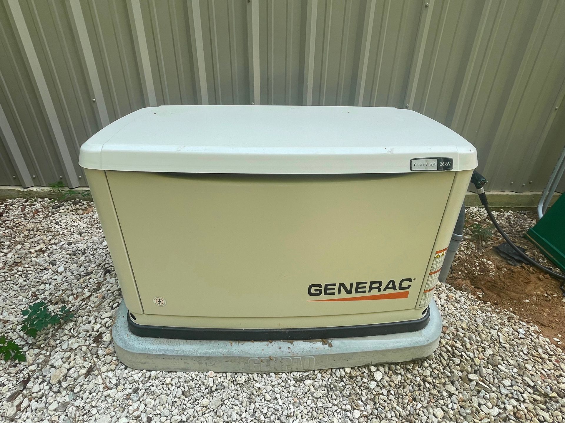 Tan Generac generator on a concrete base, set on gravel, near a corrugated metal wall.