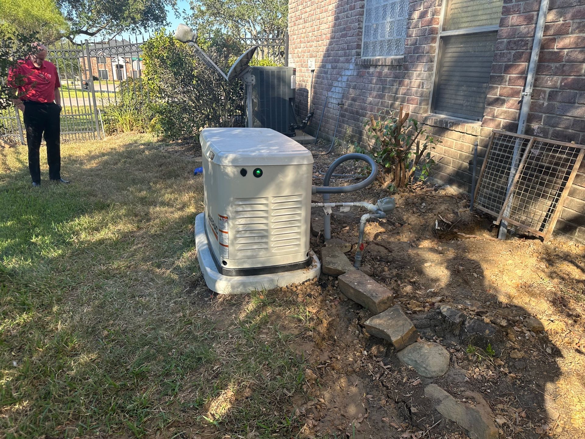 A person stands near a home generator on a grassy lawn next to a brick building.