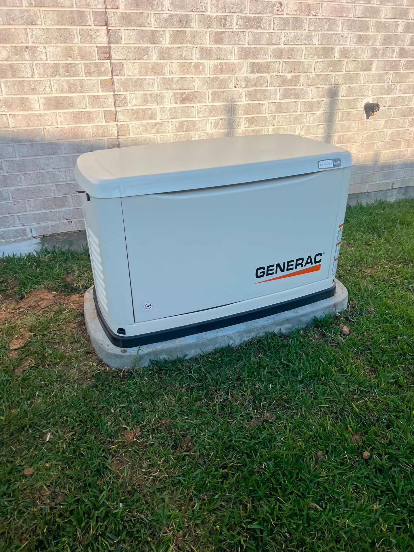 A beige Generac generator on a concrete pad, placed on grass, next to a brick wall.