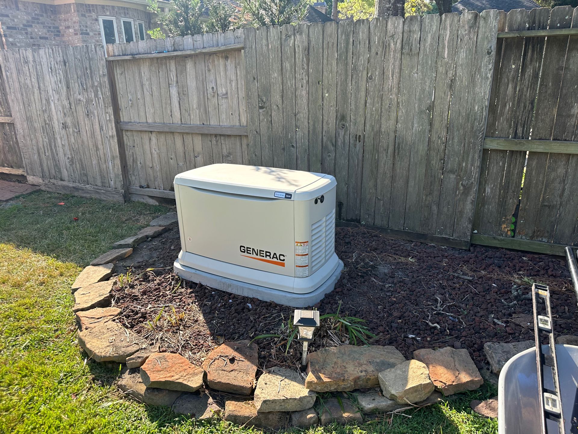 Generator near a wooden fence and garden bed. Beige and gray colors, outdoors.