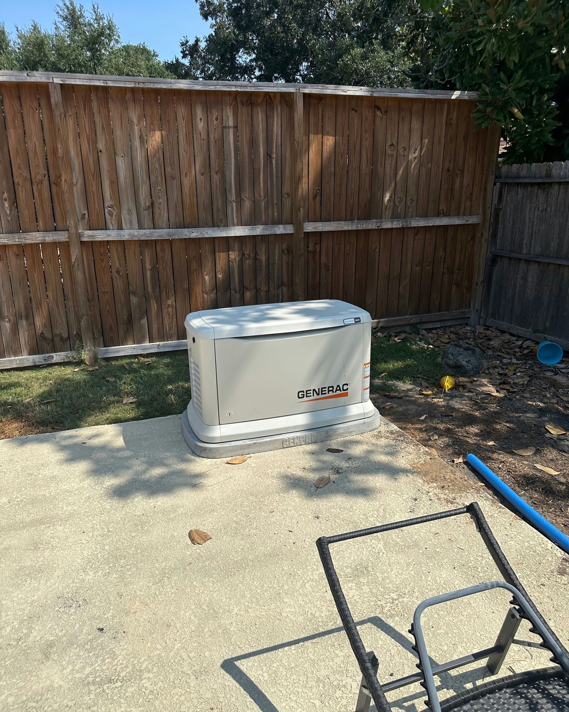 A gray generator sits on a gravel base near a wooden fence and patio.