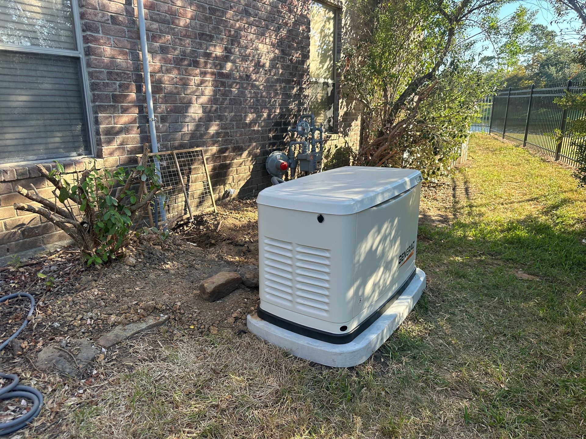 A generator sits on a concrete base next to a brick wall and a bush on a lawn.
