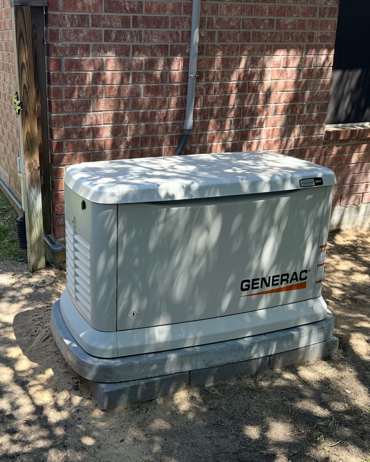 Generac standby generator, beige, on a concrete pad next to a brick wall.