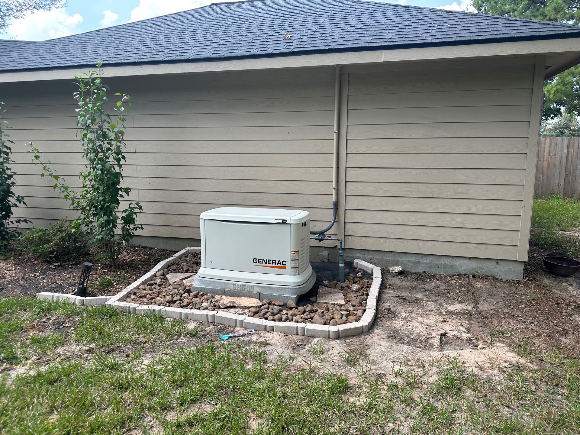 A standby generator, beige, is in a landscaped bed next to a house with tan siding and a dark roof.